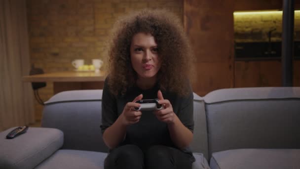 20-letnia kręcona kobieta grająca w gry wideo oglądająca przed kamerą. Podekscytowany i skoncentrowany żeński gracz z joystickiem w ręku grający w gry konsolowe. Młody dorosły udaje się w grach. — Wideo stockowe
