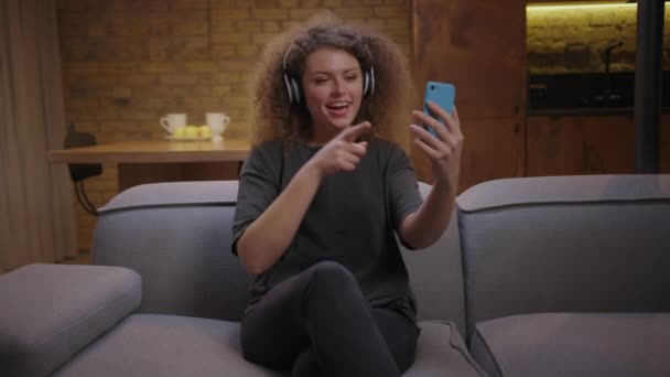 Millennial donna che canta canzoni guardando il telefono cellulare e indossando cuffie wireless. Giovane femmina gode di stare a casa seduta sul divano. — Video Stock
