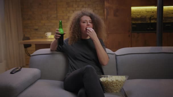 Kobieta lat 20 lubi oglądać mecz sportowy w telewizji z popcornem i piwem siedzącym na kanapie. Gry Fanka piłki nożnej lubi piłkę nożną tv gry. — Wideo stockowe