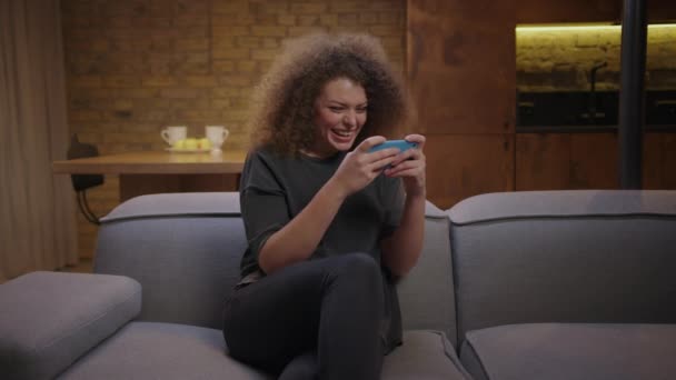 Mujer joven disfruta jugando juego de teléfono móvil sentado en el sofá en casa. 20s mujer gadget adicto utilizando el teléfono celular para juegos. — Vídeos de Stock