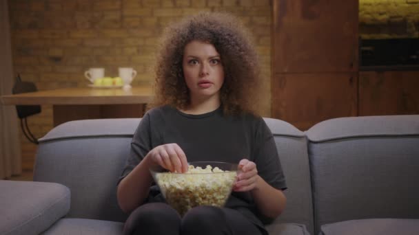 Donna anni '20 stupita guardando la TV e mangiando pop corn seduto sul divano a casa. ricci 20s donna guardando interessante film. — Video Stock