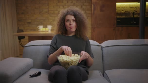 Spokojená mladá žena se dívá na televizi a jí popcorn sedící doma na gauči. Žena dívá na kameru před televizí. — Stock video