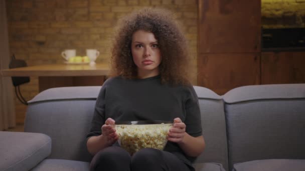 Mladá dvacetiletá žena se dívá na strašidelný film, jak sedí sama na gauči s popcornem. Vyděšená žena se dívá na televizi a zavírá oči rukama. — Stock video