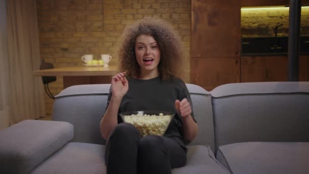 Ładna, kręcona kobieta śmiejąca się przed telewizorem w domu. Tysiąclecia kobieta ogląda film komediowy i zjada popcorn siedząc na kanapie. — Wideo stockowe