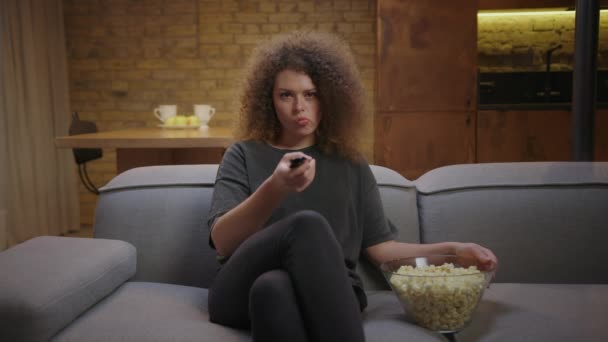 Giovane donna alla ricerca di canali TV con telecomando seduto sul divano con popcorn. riccio femmina gode guardando film con popcorn. — Video Stock