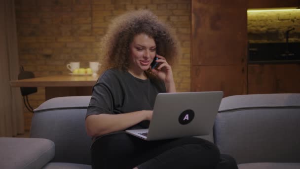 Mladá žena mluví do mobilu a dívá se na laptop, jak sedí doma na gauči. Samice, která objednává po telefonu. — Stock video