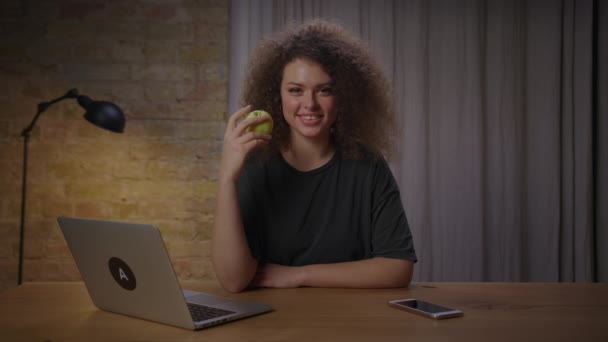 Молода жінка їсть зелене яблуко, сидячи за столом з ноутбуком вдома і дивлячись на камеру. Усміхаючись кучеряве самка любить їсти здорову їжу . — стокове відео