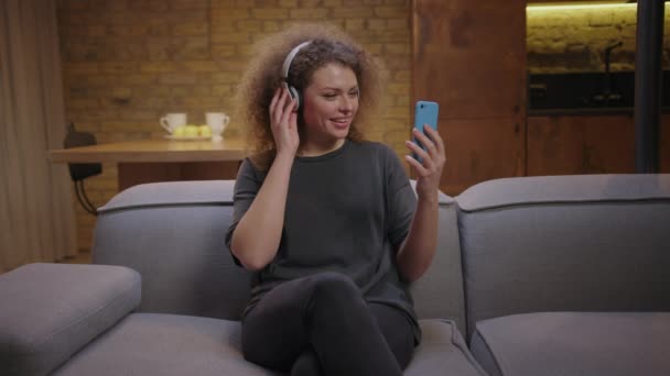 自宅のソファに座っている携帯電話を使って、ビデオ通話で話すワイヤレスヘッドフォンの20代の女性。何百万人もの女性がオンラインで両親とチャット. — ストック動画