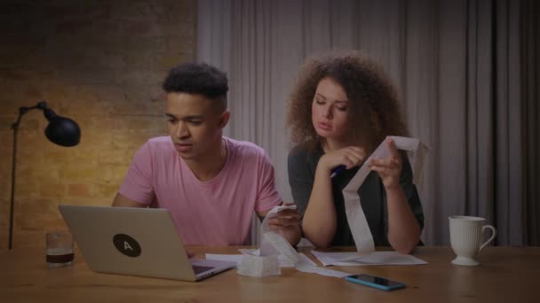 家族の予算を管理する請求書と領収書を調べる若い多民族家族。アフリカ系アメリカ人の夫は紙幣を検査する妻とラップトップで働いてコーヒーを飲む. — ストック動画