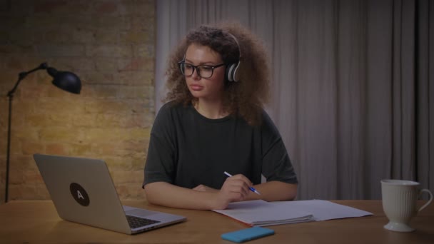 Estudiante adulto joven que tiene examen en línea en el ordenador portátil. mujer de 20 años en auriculares inalámbricos y anteojos escribiendo con pluma en papel escuchando a un profesor en línea. — Vídeo de stock