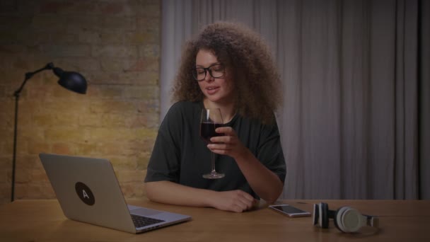Молодая 20-летняя женщина в очках пьет вино с другом онлайн на ноутбуке из-за социальной дистанции. Женщина пьет красное вино видео, звонящее домой. — стоковое видео