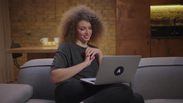 Een jonge vrouw die thuis op de bank met een laptop praat via een videogesprek. Millennial vrouw heeft online conferentie op laptop thuis. Glimlachende dame gebarentaal naar computer. — Stockvideo