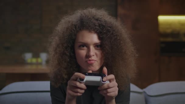 20s mulher encaracolada jogando videogame assistindo a câmera. Jogadora feminina excitada e concentrada com joystick nas mãos jogando jogos de consola. — Vídeo de Stock