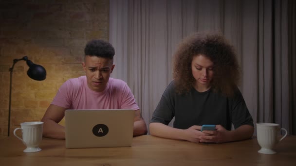 Petit ami afro-américain tapant sur ordinateur portable et petite amie bouclée surfer en ligne sur téléphone mobile. Jeune couple mixte utilisant des gadgets à la maison. Curieux homme et femme regarder les uns les autres gadgets. — Video