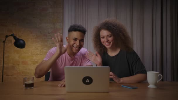Genç, çok ırklı bir çift evdeki masada dizüstü bilgisayarla konuşuyor. Afrikalı Amerikalı erkek arkadaşı ve kız arkadaşı birlikte gülüyorlar. Video aracılığıyla arkadaşlarıyla konuşuyorlar.. — Stok video