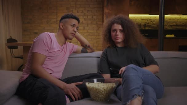 20er Jahre gemischtes Rassenpaar, das auf der Couch sitzend Film guckt und Popcorn isst. Afroamerikanisch Freund und lockige Haare Freundin vor dem Fernseher zu Hause. — Stockvideo