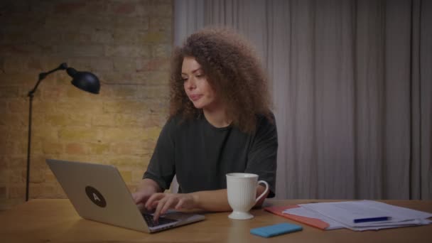 Millennial vrouw kreeg een idee om haar werk op laptop zitten aan tafel thuis af te maken. Tevreden jonge vrouw sluiten laptop en het drinken van hete thee. — Stockvideo