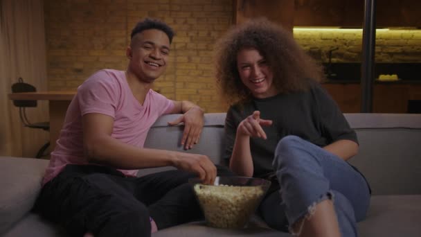 Millennial gemischtes Rassenpaar, das auf der Couch sitzend einen Comedy-Film anschaut und Popcorn isst. Schöner afroamerikanischer Freund und lockige Freundin lachen zu Hause vor dem Fernseher. — Stockvideo