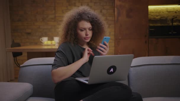 20 'li yaşlarda bir kadın evdeki kanepede oturan dizüstü bilgisayardan cep telefonunu arıyor. Genç bir bayan online alışveriş için çevrimiçi mağazayı arıyor.. — Stok video
