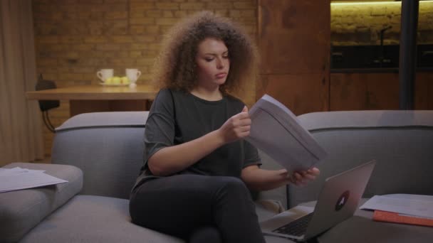 Felnőtt nő diák nézi a papíralapú dokumentumokat, és jegyzetel a laptopon ül a kanapén este otthon. Fiatal nő tanulás online otthon. — Stock videók