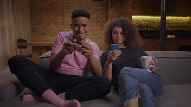 밀레니엄 혼합 인종 커플이 소파에서 저녁 시간을 함께 보내고 있습니다. 젊은 아프리카 미국 남자 비디오 게임 및 20 대 여자 온라인 서핑 휴대 전화를 사용. — 비디오