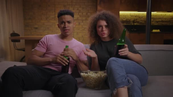 Jovem casal multiétnico assistindo jogo de futebol com pipocas e cerveja sentados no sofá. Afro-americano fã de futebol e sua namorada como o jogo de esporte de tv. — Vídeo de Stock