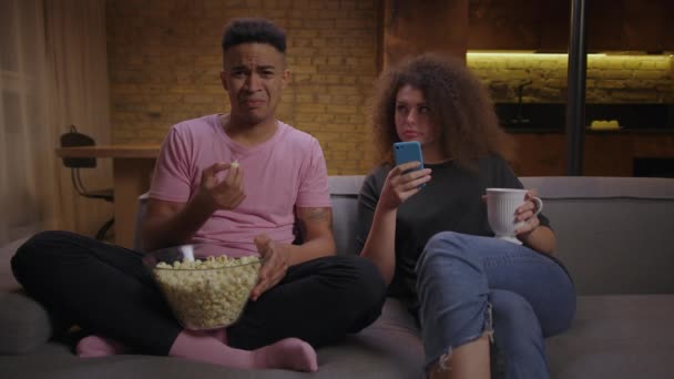 Millennial coppia di razza mista seduta sul divano la sera. 20s ragazzo afroamericano guardare la TV e mangiare pop corn, e capelli ricci ragazza navigare in internet utilizzando il telefono cellulare. — Video Stock