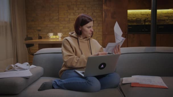 30-as évekbeli nő, aki papírokkal és laptoppal dolgozik, otthon ül a kanapén esténként. Fiatal nő távolról dolgozik otthonról. — Stock videók