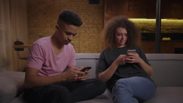 Uomo afroamericano che scrive sul cellulare e donna riccia che naviga online sul cellulare. Giovane coppia di razza mista utilizzando gadget seduti sul divano. Millennial persone phubbing a vicenda con gadget. — Video Stock