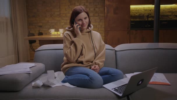 30-talskvinna som pratar via mobiltelefon sittandes på soffan med dokument på kvällen. Ung kvinna som arbetar långt hemifrån. — Stockvideo