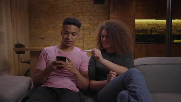 Pozitif hamilelik testi olan 20 'li yaşlarda çok ırklı bir çift. Mutlu kız arkadaş evde kanepede oturan şok olmuş erkek arkadaşına hamilelik testi gösteriyor.. — Stok video