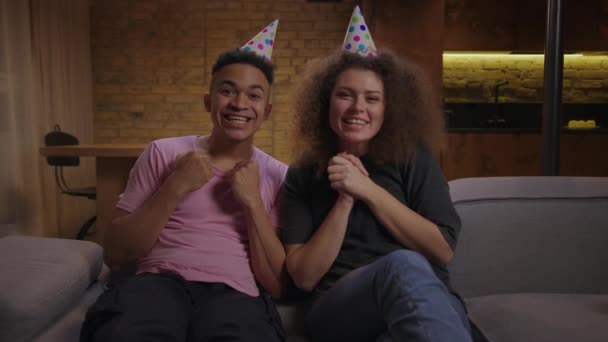 年轻的、头戴生日礼帽的混血种族夫妇坐在家里的沙发上，祝他们在摄像机前交谈愉快。网上庆祝生日的照片. — 图库视频影像