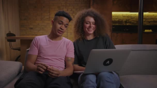 Ein multiethnisches Paar der 20er Jahre im Gespräch mit einem Laptop, der zu Hause auf der Couch sitzt. Afroamerikanischer Freund und seine Freundin lachen gemeinsam und unterhalten sich per Videoanruf mit Freunden. — Stockvideo