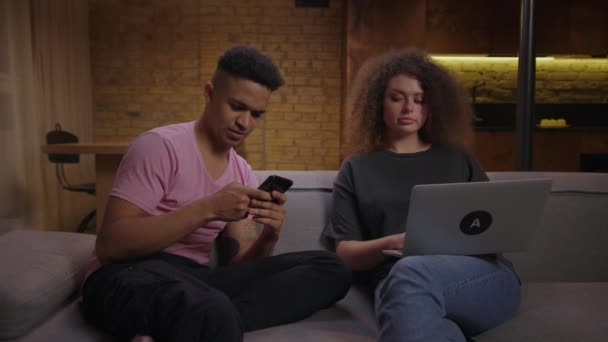 아프리카계 미국인 남자 친구가 휴대폰으로 문자 메시지를 보내고 곱슬곱슬 한 여자 친구가 소파에 앉아 노트북으로 일하고 있습니다. 여러 민족의 젊은 부부가 집에서 기기를 사용 한다. 예술가들의 물건을 보는 호기심많은 여자. — 비디오