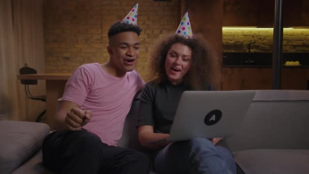 Millennial Mixed Race Paar mit Geburtstagshüten gratuliert jemandem, der mit Laptop spricht. Geburtstagsfeier online. Internet-Feier und soziale Distanz. — Stockvideo
