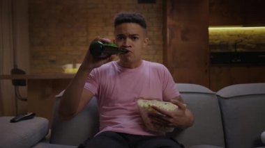 Genç, Afro-Amerikan bir adam bira ve patlamış mısırla maç seyrediyor ve kameraya bakıyor. Erkek futbol fanatiği en sevdiği takımı TV karşısında otururken destekliyor..