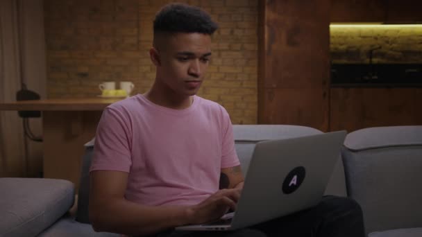 Freelancer afro-americano de 20 anos trabalhando remotamente em casa sentado no sofá. Jovem homem de trabalho concentrando digitação no laptop. — Vídeo de Stock