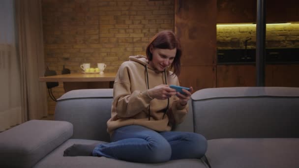 Jeune femme adulte jouant à des jeux mobiles sur téléphone mobile et gagner le jeu assis sur le canapé. Femme faisant geste de victoire avec les mains tout en jouant sur le téléphone intelligent. — Video