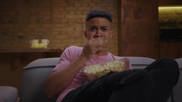 Millennial άνθρωπος βλέποντας ταινία τρόμου τρώει ποπ κορν κάθεται μόνη της στον καναπέ στο σπίτι. Φοβισμένος αρσενικό βλέποντας τηλεόραση και να τρομάξει ξαφνικά. — Αρχείο Βίντεο