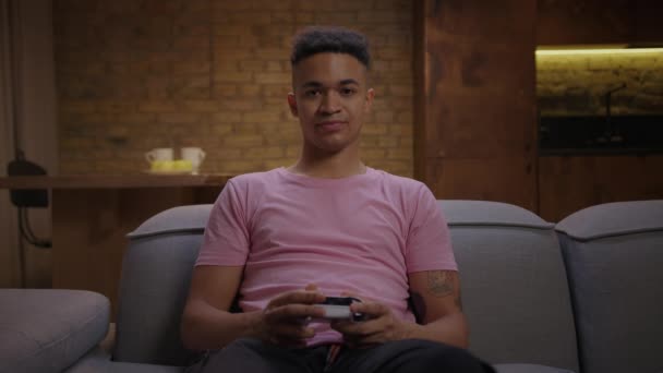 Fiatal, felnőtt afro-amerikai férfi videojátékozik a kanapén lévő kamera előtt. Unalmas férfi játék játékos joystick a kezében játszik konzol játékok. — Stock videók