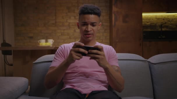 Millennial hombre afroamericano jugando videojuegos con teléfono móvil sentado en el sofá. Joven ganando el juego móvil y haciendo sí gesto. — Vídeo de stock