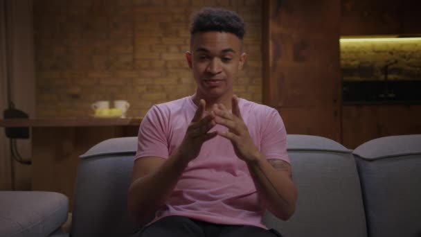 若い大人のアフリカ系アメリカ人男性のピンクのTシャツのカメラソファの上に座って話している。オンラインインタビューを受けている千年の自信のある男性。ウェブカメラビューの男の話とジェスチャー. — ストック動画