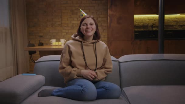 Joven mujer alegre de 30 años con sombrero de cumpleaños felicitando con cumpleaños hablando en la cámara sentado en el sofá. Felicitaciones de cumpleaños en línea. — Vídeo de stock