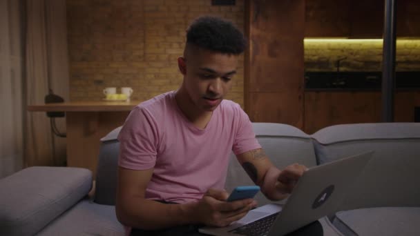 20 대 아프리카계 미국인이 소파에 앉아 온라인 쇼핑을 하기 위해 전화를 걸었습니다. 기꺼이 집에서 온라인으로 물건을 사려는 젊은 남자. — 비디오