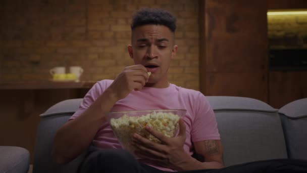 Pembe tişörtlü, patlamış mısır yiyen ve evdeki kanepede oturan genç Afro-Amerikan bir adam. Genç yetişkin heyecanlı bir film izliyor.. — Stok video