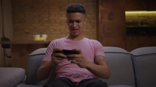 Νεαρός Αφροαμερικανός παίζει κινητό παιχνίδι στο smartphone κάθεται στον καναπέ. Συγκεντρωμένος παίκτης χιλιετηρίδας χρησιμοποιώντας το κινητό τηλέφωνο για online gaming. 20s άνθρωπος γιορτάζει τη νίκη στο κινητό εφαρμογή. — Αρχείο Βίντεο