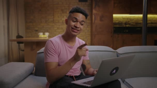 若いアフリカ系アメリカ人の男性は、自宅のソファに一人で座っているビデオ通話でラップトップに話して笑っています。社会的距離通信. — ストック動画