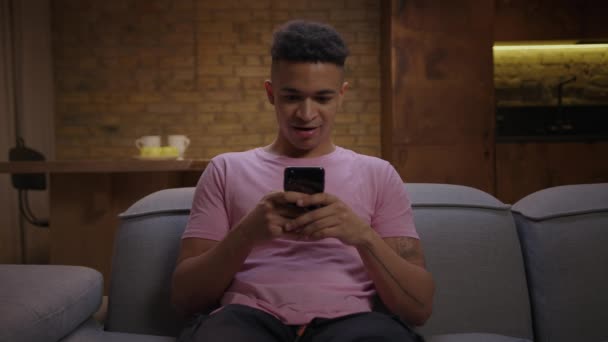 Millennial Αφρικής Αμερικανός άνθρωπος πήρε ενθουσιασμένος χρησιμοποιώντας το κινητό τηλέφωνο κάθεται στον καναπέ στο σπίτι. Νεαρός διαβάζει καλά νέα και προκαλεί έκπληξη στο πρόσωπό του.. — Αρχείο Βίντεο