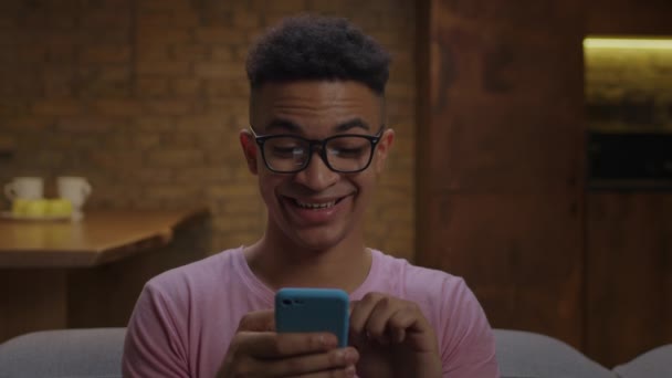 Hombre afroamericano de 20 años usando anteojos usando teléfono celular y navegando por internet sentado en el sofá en casa. Primer plano de gadget adicto joven masculino de navegación en la red social. — Vídeos de Stock