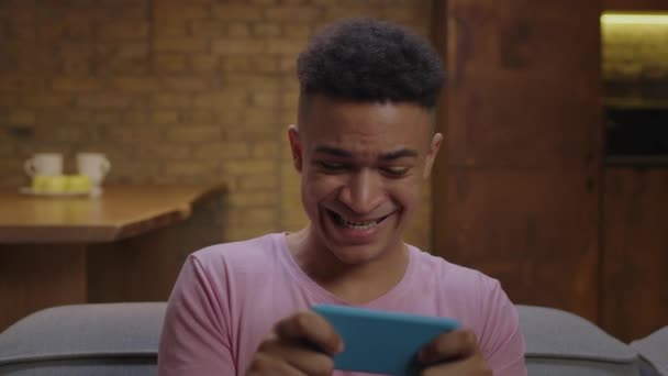 Millennial homem afro-americano jogando videogames usando telefone celular sentado no sofá. Close up de jovem ganhando o jogo móvel e fazendo sim gesto de vitória. — Vídeo de Stock
